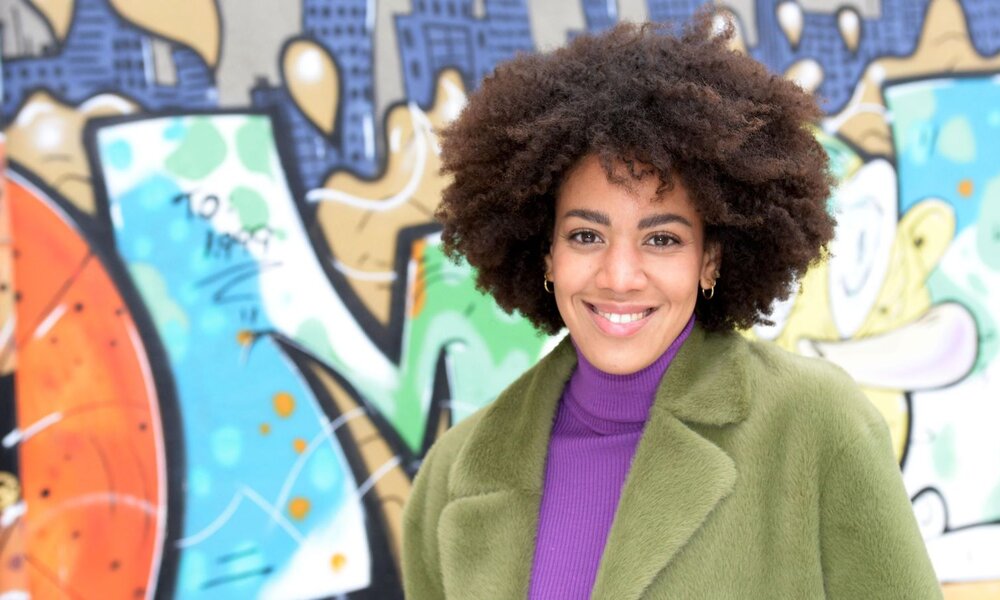 Eine fröhliche Frau mit dunklen Locken und goldenen Ohrringen posiert vor einer farbenfrohen Graffiti-Wand in der Stadt. | © RTL / Rolf Baumgartner