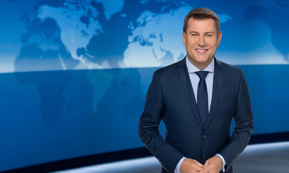 Ein lächelnder Nachrichtensprecher im dunkelblauen Anzug steht vor einer blauen Weltkarten-Grafik im TV-Studio. | © NDR / Thorsten Jander