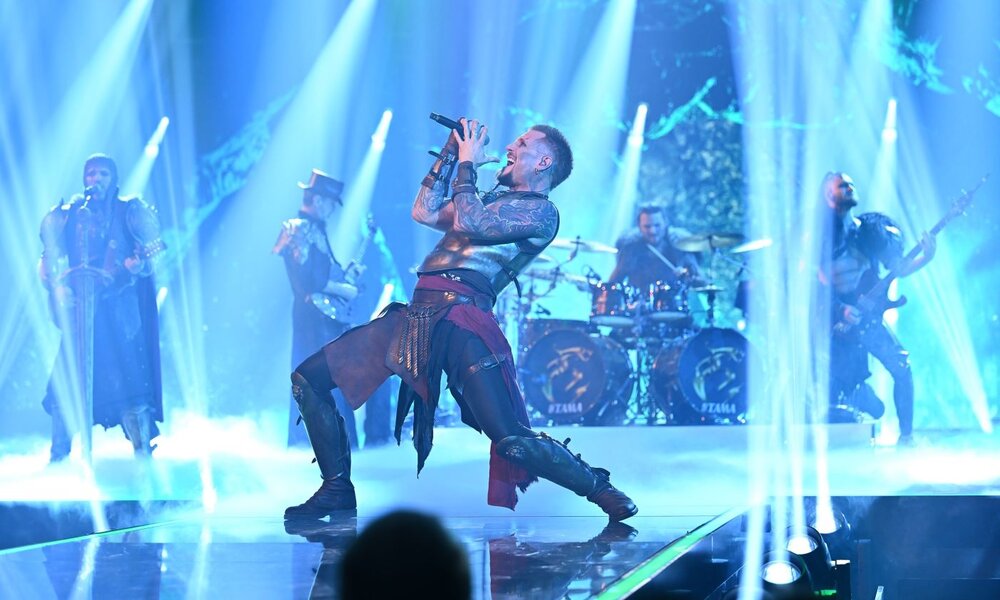 Eine Metal-Band in mittelalterlicher Kriegsrüstung performt energiegeladen auf einer nebligen Bühne, während der Sänger dramatisch ins Mikrofon singt. | © NDR/Raab ENTERTAINMENT/Willi Weber