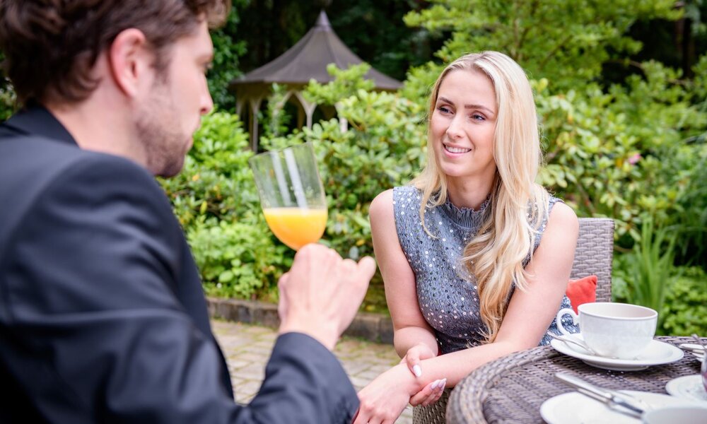 Ein Mann mit dunklen Haaren hält ein Glas Orangensaft, während eine blonde Frau ihm lächelnd zuhört, umgeben von grüner Natur. | © ARD/WDR/Christof Arnold 