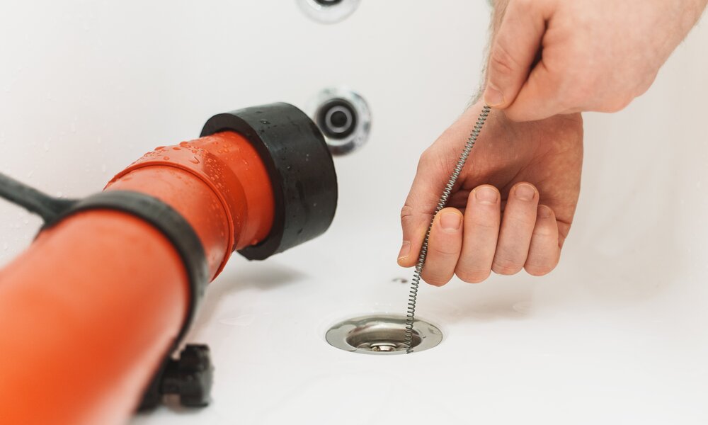 Nahaufnahme einer Rohrreinigung: Eine Hand hält eine Abflussspirale, die in einen verchromten Wannenabfluss eingeführt wird, neben einem feuchten Rohr. | © Adobe Stock/ M-Production