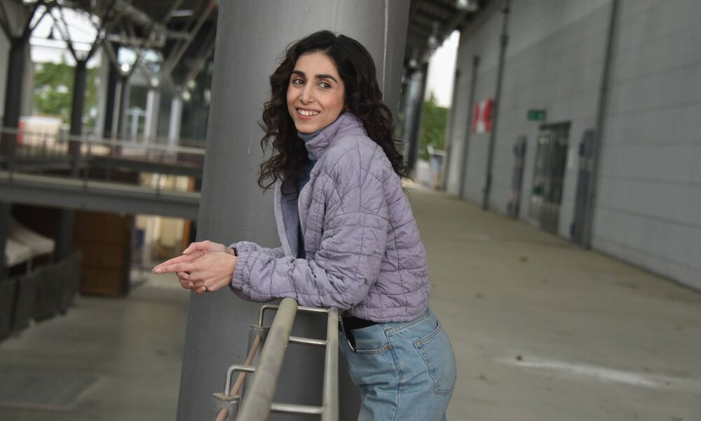Eine junge Frau mit dunklen Locken lehnt lächelnd an einem Geländer, trägt eine lila Steppjacke und Jeans in einer modernen Industriehalle. | © IMAGO Images/ Horst Galuschka