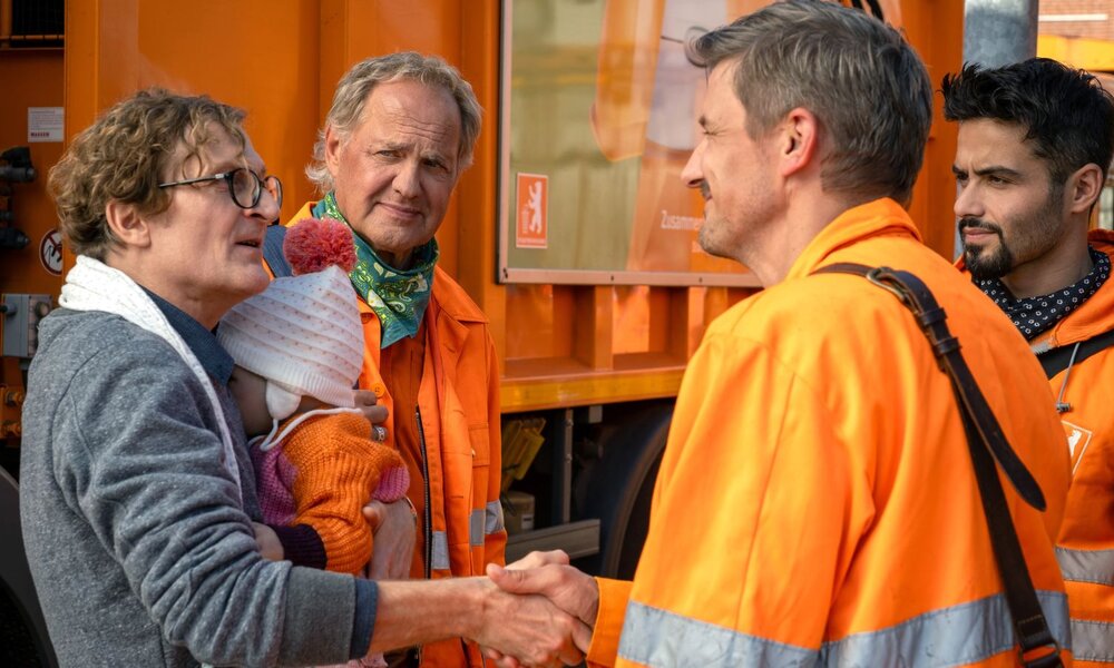 Ein Mann mit Brille und grauem Pullover hält ein Kleinkind in bunter Winterkleidung im Arm und schüttelt einem Müllwerker die Hand. | © ARD Degeto Film/Conny Klein