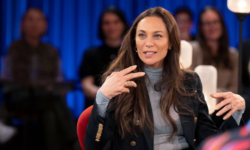 Eine braunhaarige Frau in dunklem Blazer und grauem Rollkragenpullover gestikuliert lächelnd während einer Talkshow vor blauem Hintergrund. | © WDR/Melanie Grande