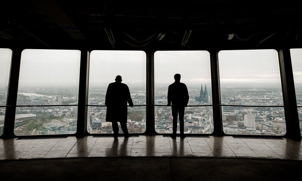 Zwei Männer stehen als Silhouetten vor großen Fenstern in einer Aussichtsplattform und blicken auf die Kölner Skyline mit Dom. | © WDR/Sandra Stein