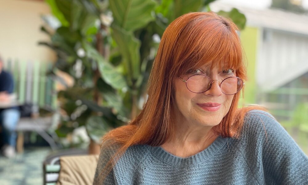 Eine rothaarige Frau mit Brille und blauem Strickpullover lächelt sanft in die Kamera, während sie in einem Café mit grünen Pflanzen sitzt. | © BR/Evelyn Schels