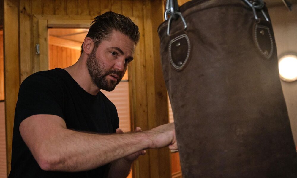 Ein bärtiger Mann mit dunklem, zurückgekämmtem Haar und schwarzem T-Shirt trainiert konzentriert an einem schweren Boxsack in einem holzvertäfelten Raum. | © RTL / Stefan Behrens