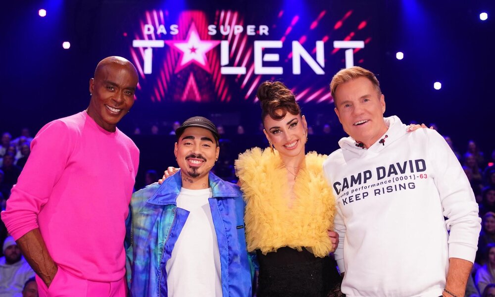 Eine Jury-Gruppe mit zwei Männern und einer Frau posiert fröhlich auf der Bühne von "Das Supertalent", umgeben von Publikum im Hintergrund. | © RTL / Stefan Gregorowius