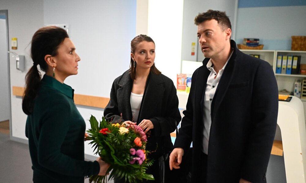 Eine dunkelhaarige Frau mit strengem Blick hält einen Blumenstrauß, während eine junge Frau und ein Mann in dunklen Mänteln sie ernst ansehen. | © RTL / Julia Feldhagen