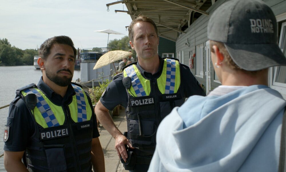 Die Polizisten Malik Hakim (Zejhun Demirov) und Mattes Seeler (Matthias Schloo) stehen nebeneinander vor Wohncontainern. Beide blicken zu Colin (Aris Wolf), der seitlich vor ihnen steht.  | © ZDF/Christian Klopp