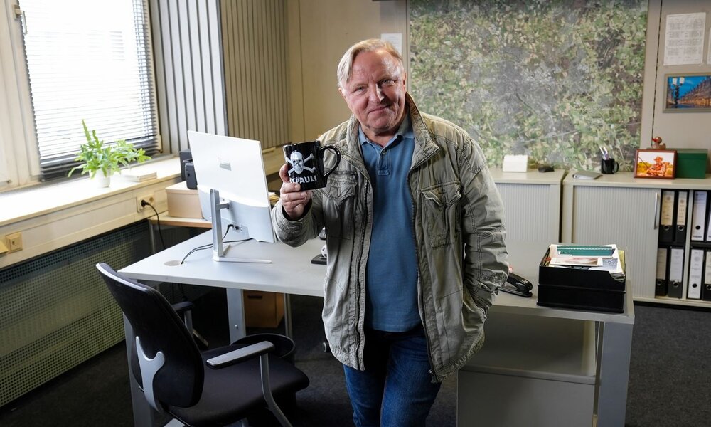 In einem Büro mit Landkarte an der Wand steht ein Mann in Jeans und grüner Jacke, lächelt und zeigt eine Tasse mit Totenkopf-Design. | © NDR/WDR/Dokfilm/Jan Urbanski