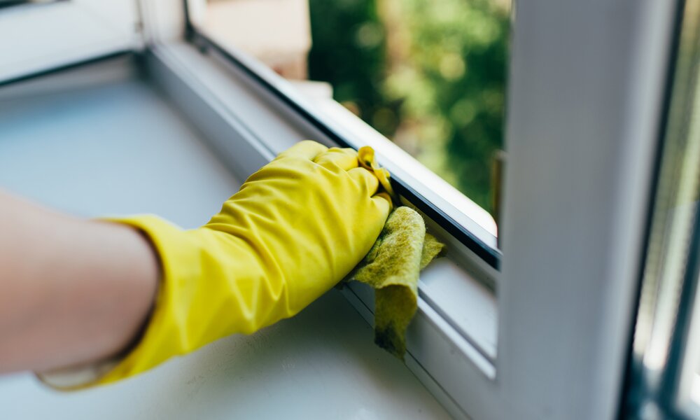 Eine Person mit gelbem Gummihandschuh reinigt mit einem feuchten Tuch den weißen Fensterrahmen. | © Adobe Stock/ gesrey
