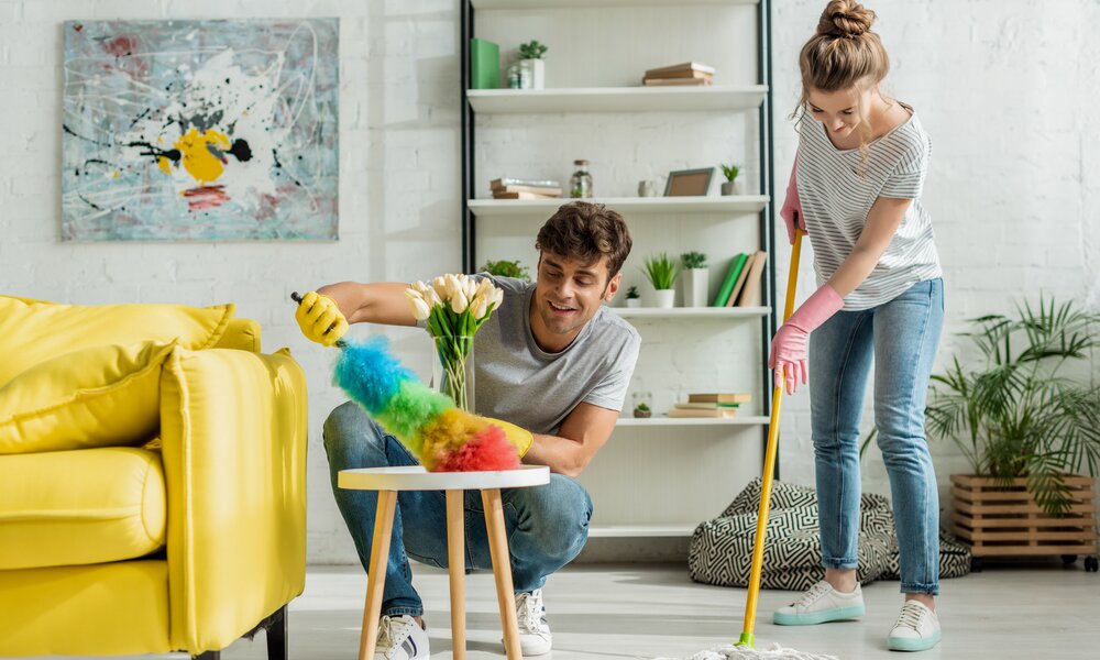 Junges Paar putzt gemeinsam lachend das Wohnzimmer, er entstaubt den Tisch, sie wischt den Boden mit einem Wischmopp. | © Adobe Stock/ LIGHTFIELD STUDIOS
