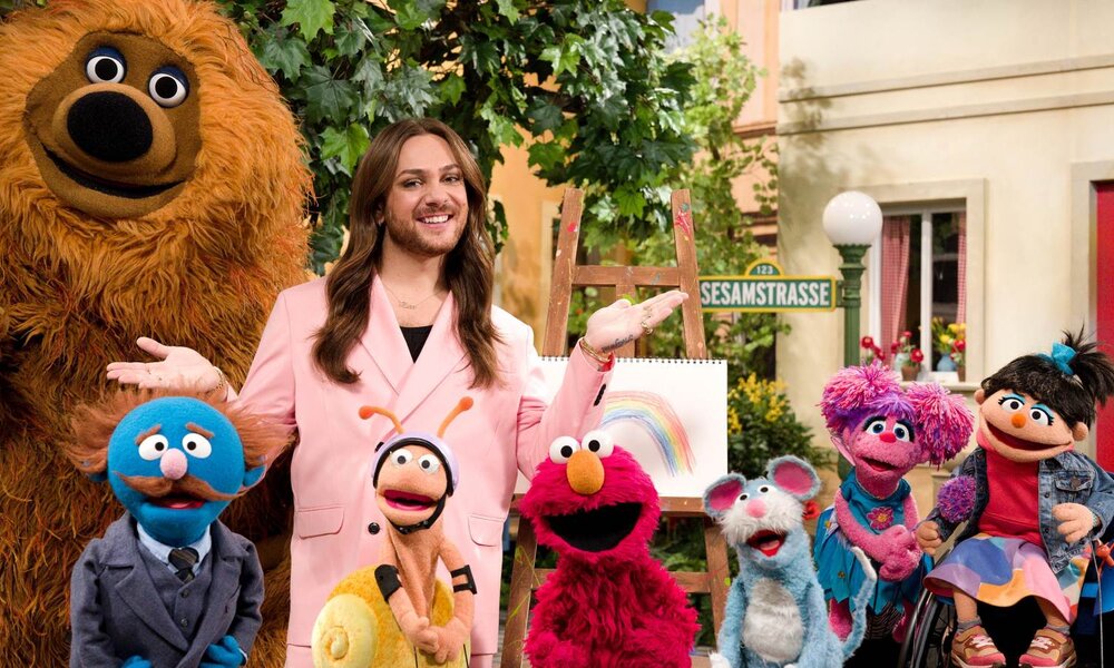 Ein junger Mann mit langen braunen Haaren in rosa Blazer steht fröhlich lächelnd mit Sesamstraßen-Figuren vor einem bunten Haus. | © NDR/Sesame Workshop/Jonathan Friesicke 