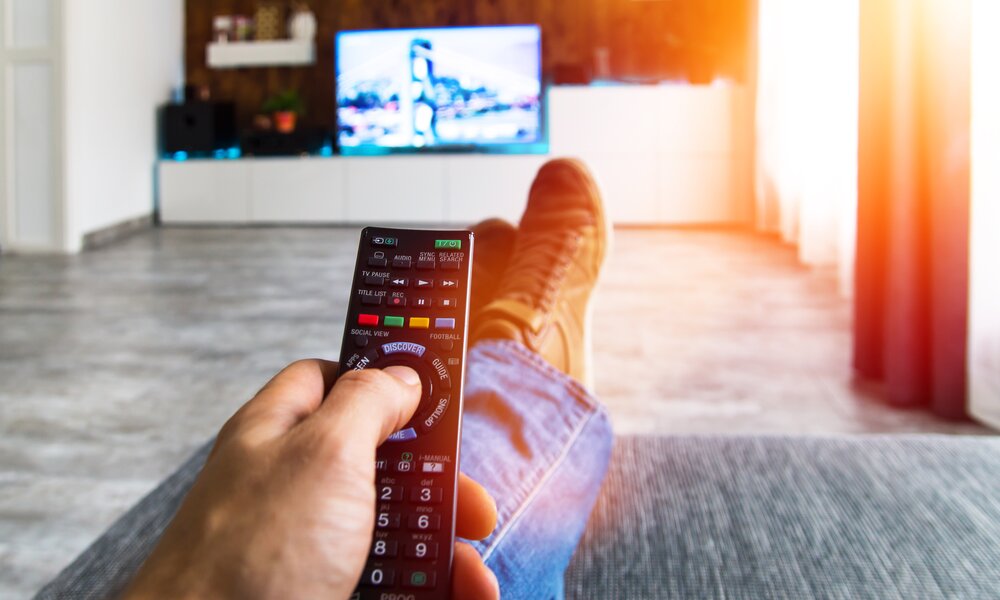 Mann sitzt auf Couch mit Fernbedienung in der Hand und vorne an Wand Fernseher an | © Adobe Stock/ghazii