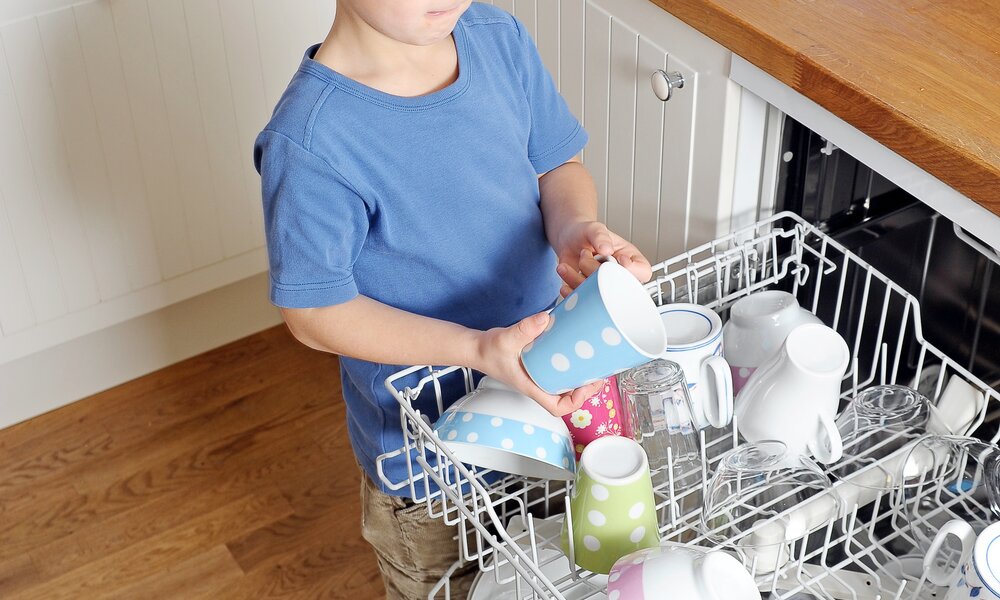 Ein kleiner Junge mit blauem Shirt steht an voller Spülmaschine und hält blaue Tasse mit Punkten | © Adobe Stock/photophonie