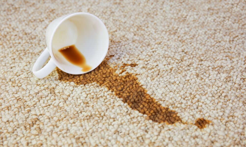Umgekippte weiße Kaffeetasse mit braunem Fleck auf hellem Teppich mit grober Struktur. | © Adobe Stock/ Viktoriia M