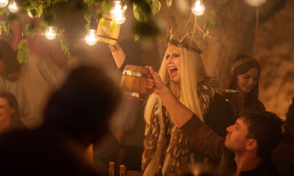 In stimmungsvoll beleuchtetem Raum hebt eine kostümierte Frau mit langem blondem Haar und Trinkhorn jubelnd ihren Becher. | © ARD Degeto Film/Odeon Fiction GmbH/Karolina Grabowska