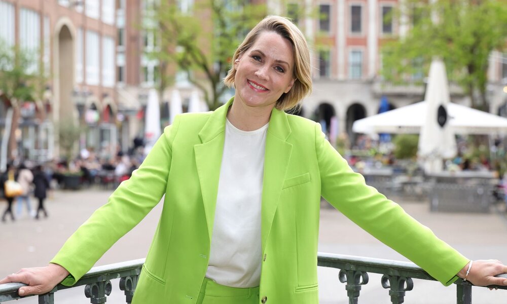 Fröhliche Frau in leuchtend grünem Hosenanzug steht mit ausgestreckten Armen auf einem Platz mit Cafés und Altbauten im Frühling. | © Getty Images Enterteinment / Andreas Rentz / Staff