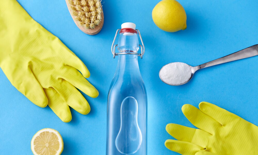 Eine Glasflasche mit Essigessenz steht umgeben von Zitronen, Natron, Bürste und gelben Gummihandschuhen auf blauem Untergrund. | © Adobe Stock/ Syda Productions