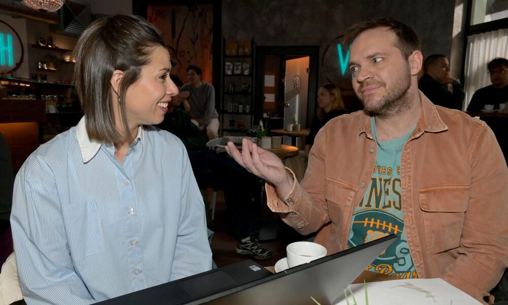 Junge Frau mit halblangem braunem Haar und hellblauer Bluse lacht einem Mann im Café zu, der mit ironischem Blick die Hand hebt. | © RTL / Rolf Baumgartner