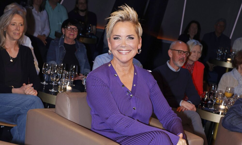 Strahlende Frau mit kurzem, blondem Haar in lilafarbenem Outfit sitzt vor einem Publikum an kleinen Tischen mit Weingläsern. | © IMAGO / Future Image / H. Hartmann