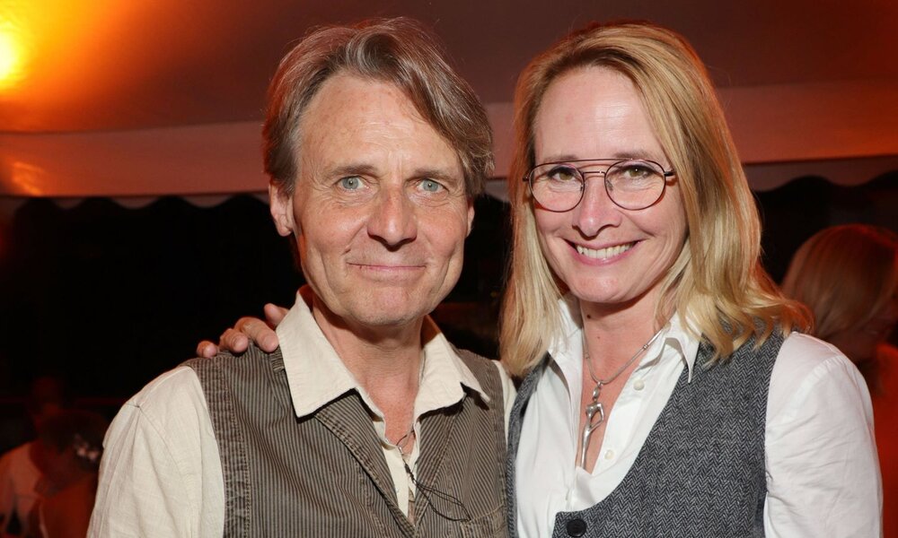 Eine blonde Frau mit Brille und ein grauhaariger Mann lächeln nebeneinander vor dunklem Hintergrund unter orangefarbenem Licht. | © IMAGO / Eventpress Becher