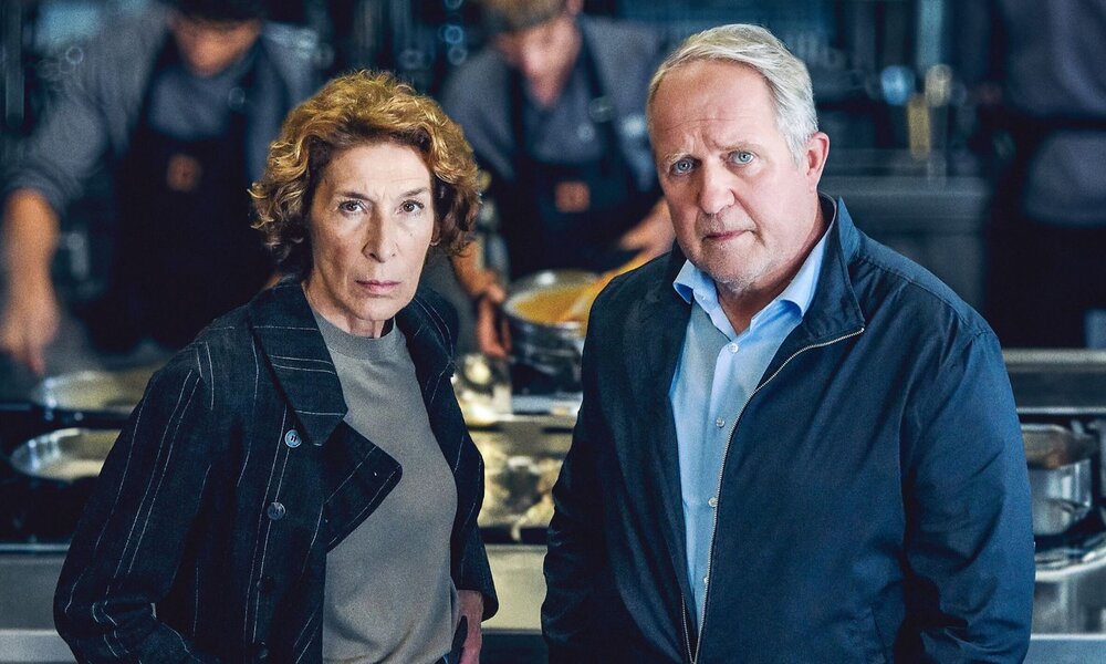 Grauhaariger Mann und Frau mit lockigem Haar blicken ernst in die Kamera, hinter ihnen geschäftiges Treiben in einer Restaurantküche. | © ARD Degeto Film/ORF/Hubert Mican