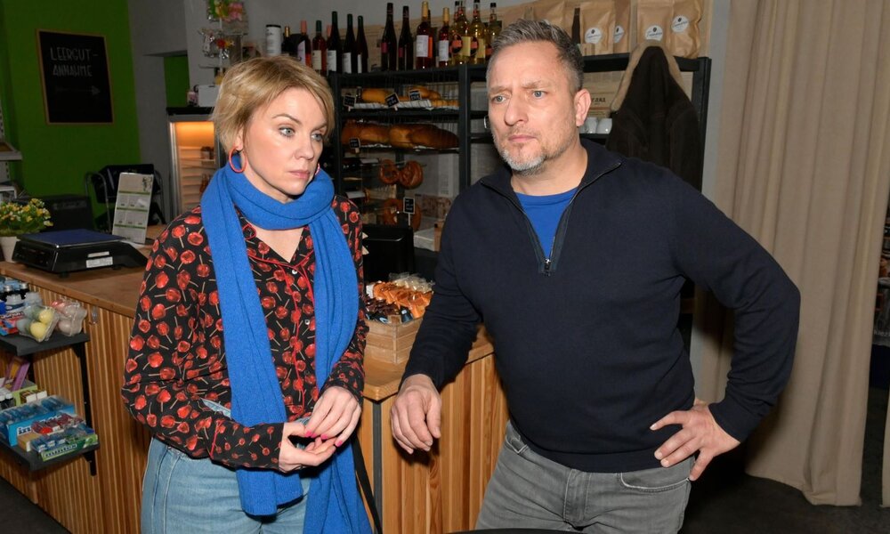 Blonde Frau mit blauem Schal und ernster Miene steht neben nachdenklichem Mann in dunklem Pullover hinter einem Tresen. | © RTL / Rolf Baumgartner