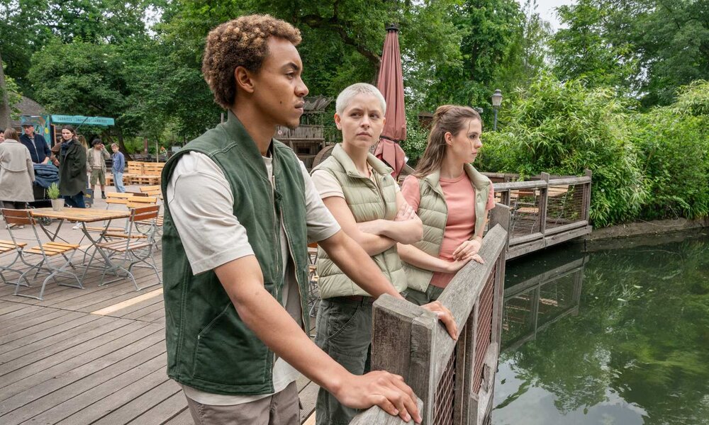 Drei junge Erwachsene in Outdoorkleidung lehnen nachdenklich am Holzgeländer eines Teichs in grüner Parklandschaft. | © ARD/Saxonia Media/Steffen Junghans 