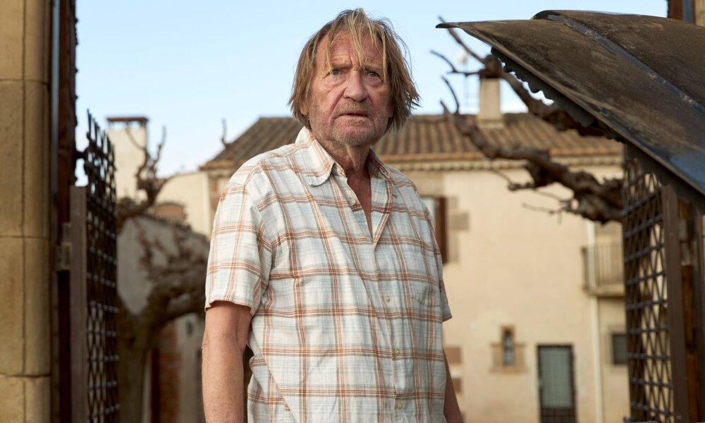 Ein älterer Mann mit zerzaustem Haar und kariertem Kurzarmhemd blickt ernst in die Kamera, umgeben von mediterraner Architektur. | © ARD Degeto Film/Lucia Faraig