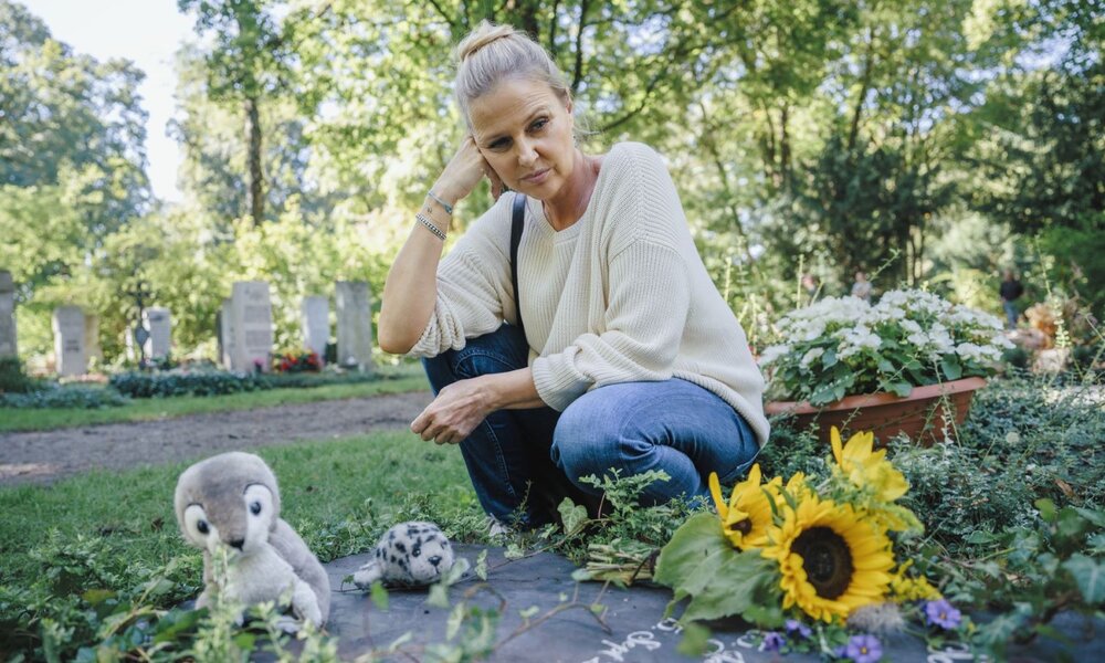 Nachdenkliche, blondhaarige Frau kniet in Jeans und cremefarbenem Pullover an einem Grab mit Sonnenblumen und Kuscheltieren. | © ZDF / Hendrik Heiden