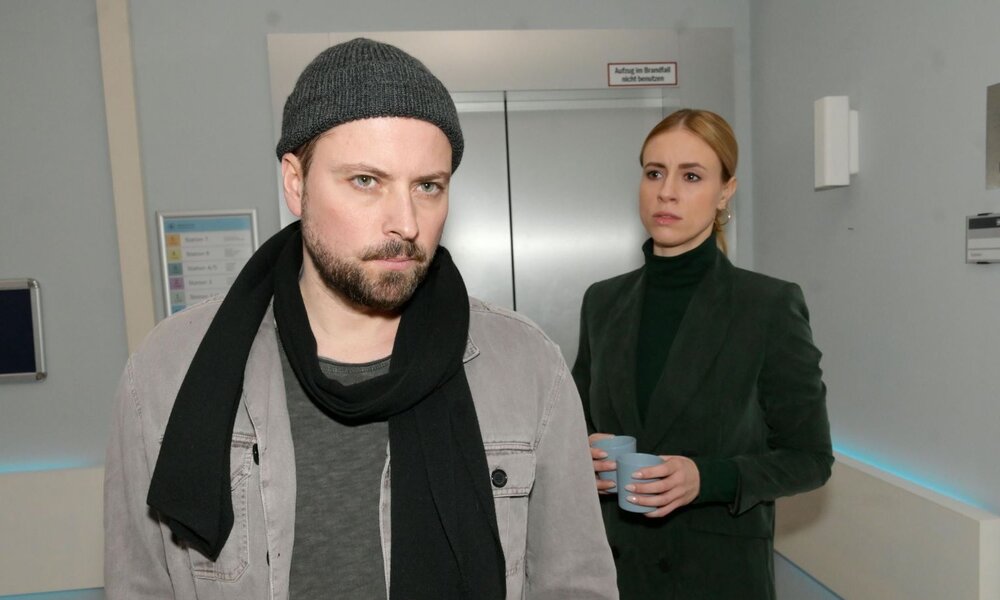 Ein bärtiger Mann mit Mütze und Schal blickt ernst, während eine blonde Frau im dunklen Blazer mit zwei Bechern hinter ihm steht. | © RTL / Rolf Baumgartner
