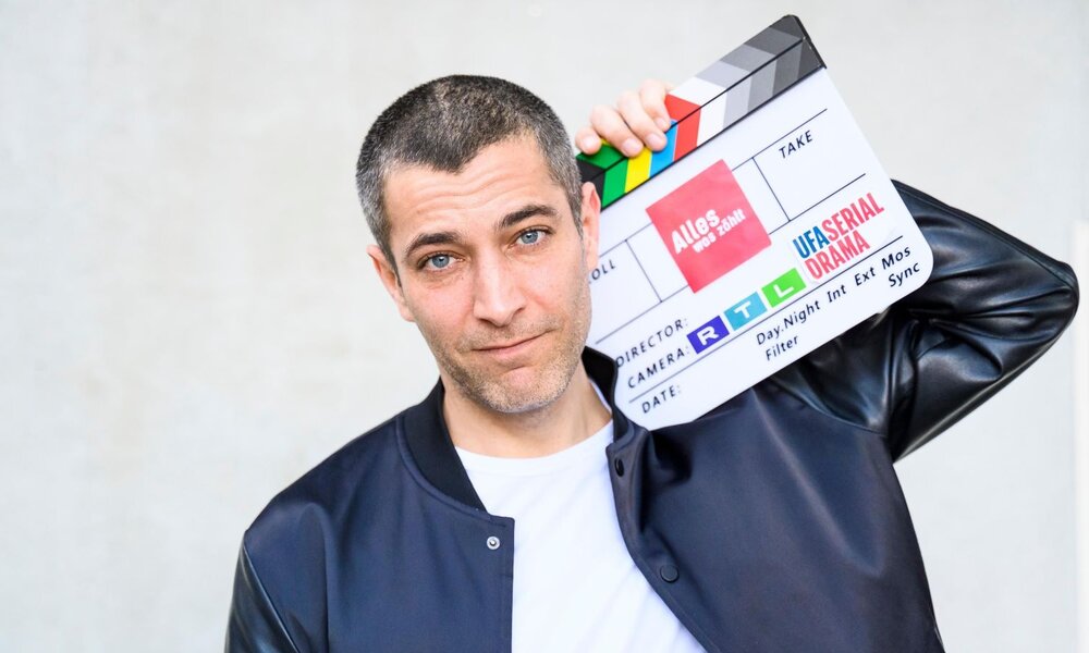 Junger Mann mit kurzen dunklen Haaren hält lässig eine Filmklappe an den Kopf, trägt weiße T-Shirt und schwarze Jacke. | © RTL / Julia Feldhagen