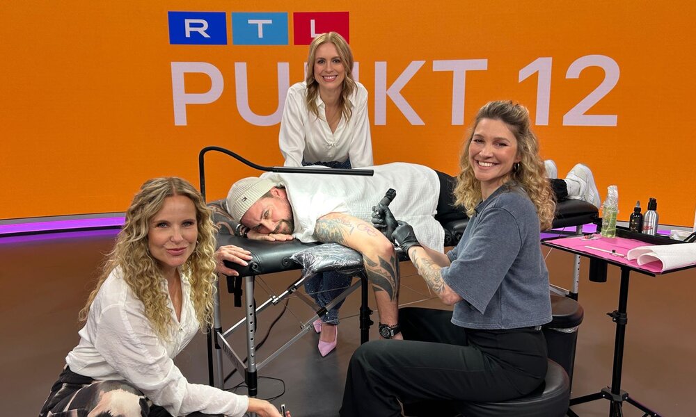 Zwei blondhaarige Frauen lächeln neben einem tätowierten Mann auf einer Liege, während eine junge Tätowiererin fröhlich arbeitet. | © RTL – Judith Pamme