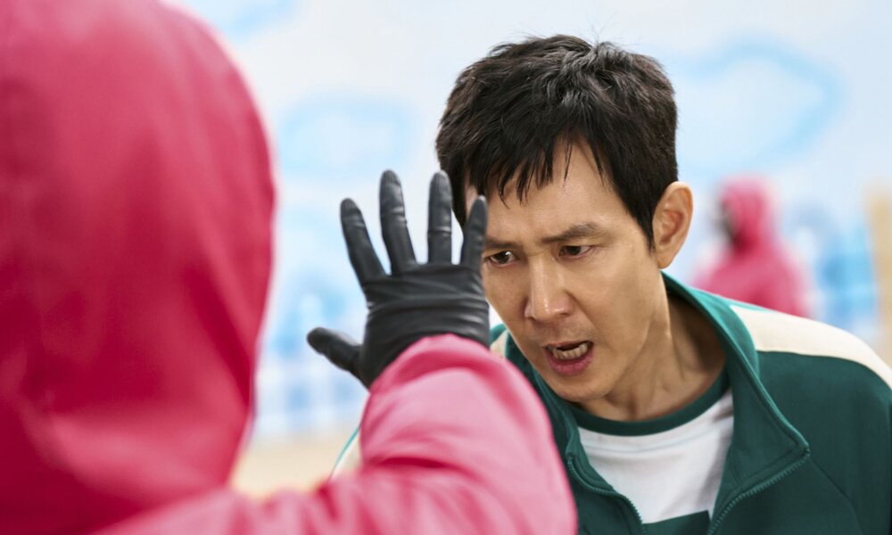 Szene aus Staffel 2 von „Squid Game“: Spieler 456 (Lee Jung-jae) schreit einen maskierten Wächter mit erhobener Hand an. | © Netflix