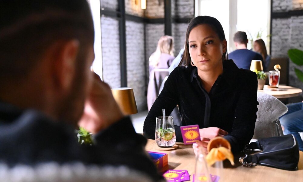 Zwei Menschen spielen an einem Holztisch ein Fragespiel in gemütlicher Bar, Frau blickt ruhig und konzentriert auf ihren Gegenüber. | © RTL / Julia Feldhagen