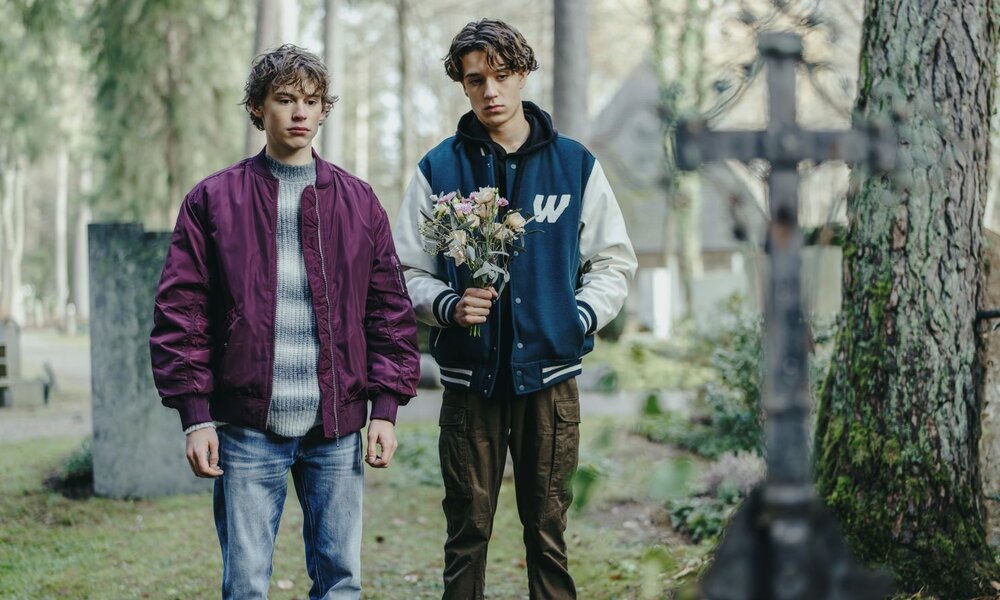 Zwei junge Männer mit lockigem Haar stehen ernst auf einem Friedhof; einer hält verblasste Blumen, hinter ihnen Bäume und Grabsteine. | © ARD Degeto Film/Bantry Bay Productions GmbH/Hendrik Heiden