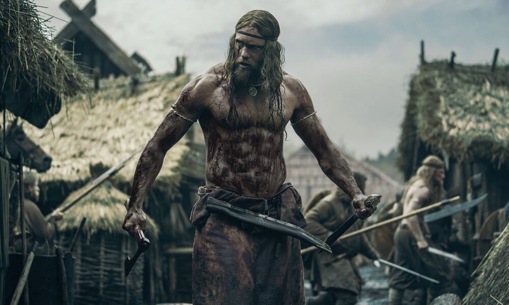 Muskulöser, blutverschmierter Mann (Skarsgård ) mit langen Haaren und Axt steht kampfbereit in einem matschigen Wikingerdorf mit Strohhütten. | © IMAGO / Capital Pictures