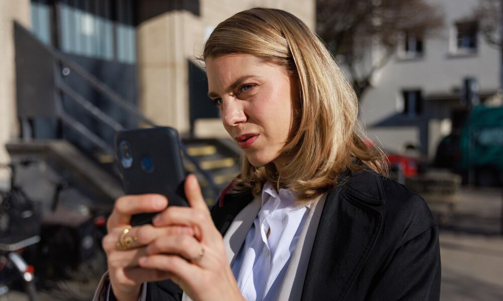 Junge blonde Frau in weißer Bluse und schwarzem Mantel hält draußen konzentriert ihr Smartphone vor einem Gebäude. | © RTL / Sebastian Meyer