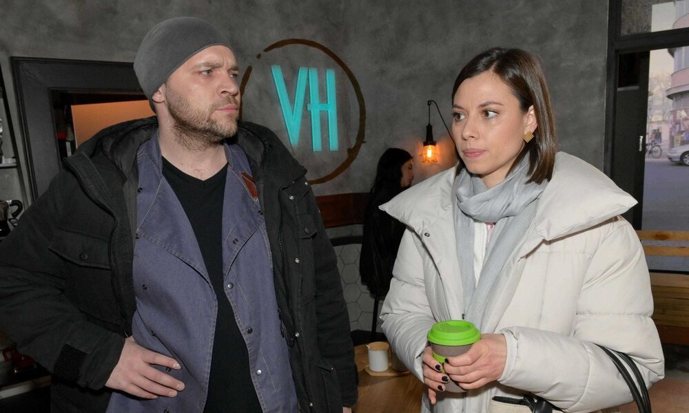 Ein bärtiger Mann in dunkler Jacke und grauer Mütze blickt skeptisch auf eine junge Frau im weißen Mantel mit Kaffeebecher. | © RTL / Rolf Baumgartner