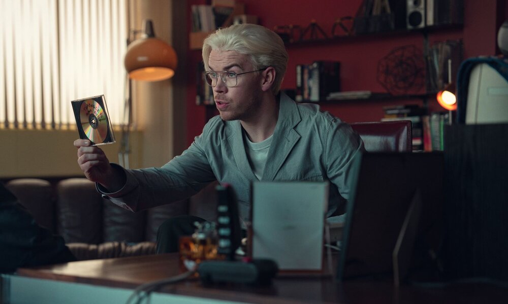 Junger Mann mit platinblondem Haar und Brille hält in einem Retro-Büro aufmerksam eine beschriftete CD in der Hand. | © Netflix