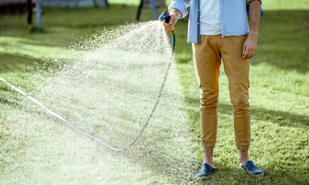 Ein junger Mann mit blauem Hemd und senffarbener Hose spritzt mit einem Gartenschlauch Wasser auf eine sonnige Rasenfläche. | © Adobe Stock/ rh2010