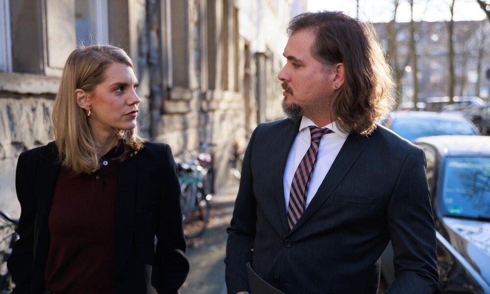 Eine junge Frau mit blondem Haar und ein Mann mit langen braunen Haaren im Anzug blicken sich ernst auf einer Straße an. | © RTL / Sebastian Meyer