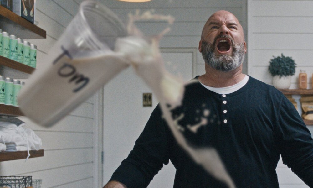 Wütender Mann mit grauem Vollbart und dunklem Shirt steht schreiend in hellem Raum, während ein beschrifteter Becher in Zeitlupe fällt. | © Netflix