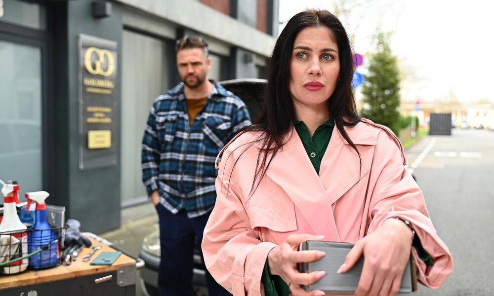 Junge Frau mit langen dunklen Haaren und rosa Mantel blickt ernst, während sie ein Paket hält; im Hintergrund ein Mann vor Werkstatt. | © RTL / Julia Feldhagen