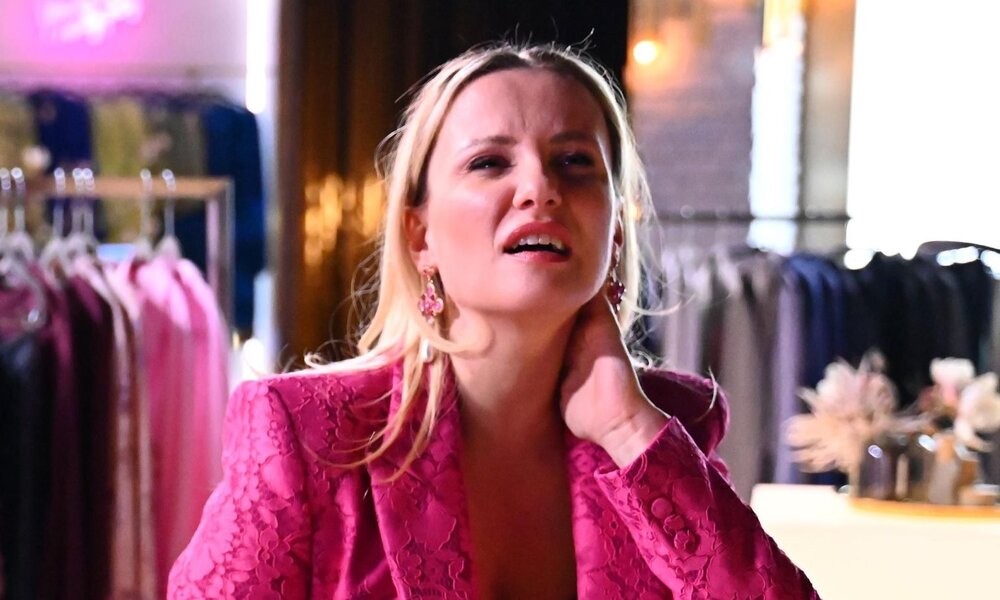 Blondhaarige Frau in auffälligem pinkem Blazer hält sich den Nacken und blickt leidend, im Hintergrund ein Modegeschäft mit Blazern. | © RTL / Julia Feldhagen
