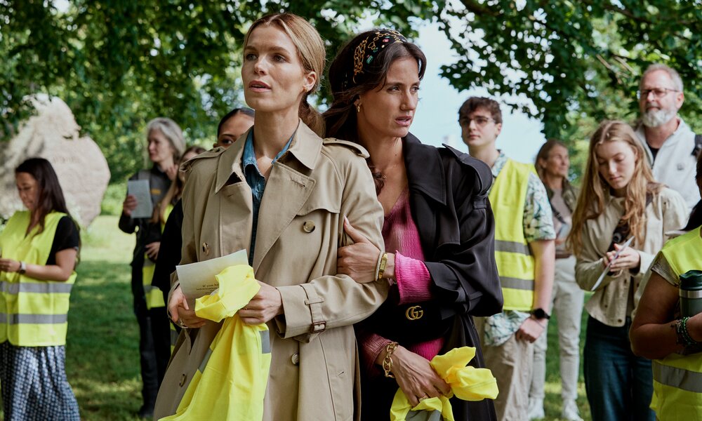 Junge Frau im Trenchcoat und Frau mit schwarzem Mantel und Stirnband halten gelbe Westen, umgeben von Menschen mit Notizen. | © Netflix