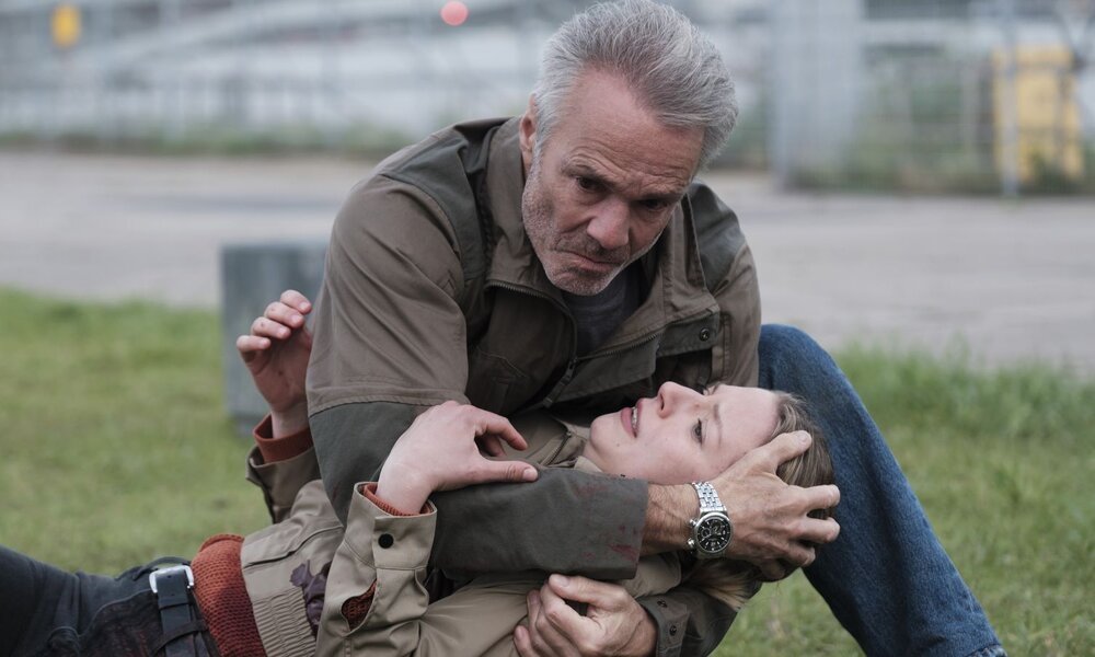Ein grauhaariger Mann kniet auf Gras und stützt eine bewusstlose Frau in beiger Jacke, Blick voller Angst und Trauer. | © ARD Degeto Film/Dinand van der Wal
