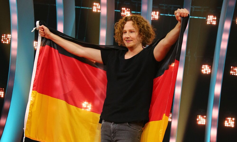 Junger Mann mit schwarzem T-Shirt und lockigem Haar breitet auf einer Showbühne die deutsche Flagge aus. | © 2018 Getty Images/Christian Marquardt